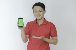 un joven asiático inteligente está feliz y sonriendo cuando muestra la pantalla verde del teléfono inteligente en el fondo del estudio foto