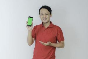 un joven asiático inteligente está feliz y sonriendo cuando muestra la pantalla verde del teléfono inteligente en el fondo del estudio foto