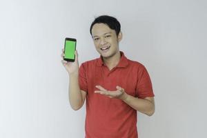 un joven asiático inteligente está feliz y sonriendo cuando muestra la pantalla verde del teléfono inteligente en el fondo del estudio foto