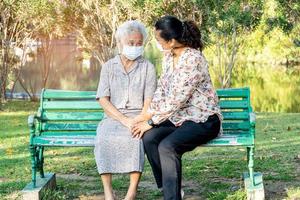 Caregiver help and talk with Asian senior or elderly old lady woman wearing a face mask for protect safety infection Covid19 Coronavirus in park. photo