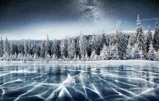 Blue ice and cracks on the surface of the ice. Frozen lake in winter mountains. It is snowing. The hills of pines. Winter. Carpathian Ukraine Europe photo