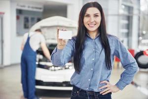 A beautiful woman keeps a business card of the car service center. The mechanic inspects the car under the hood in the background photo