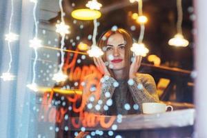 hermosa mujer joven sentada en la cafetería, bebiendo café. modelo escuchando música. navidad, feliz año nuevo, día de san valentín, concepto de vacaciones de invierno foto