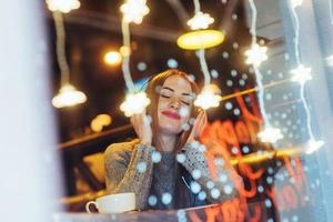 hermosa mujer joven sentada en la cafetería, bebiendo café. modelo escuchando música. navidad, año nuevo, día de san valentín, concepto de vacaciones de invierno foto