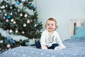 muy agradable y encantadora niñita rubia de blanco sentada en la cama y mirando la foto en el fondo de árboles de Navidad sonrientes en el luminoso interior de la casa