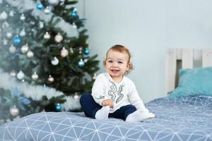 Very nice charming little girl blonde in white sitting on the bed and looking at the picture on the background of smiling Christmas trees in bright interior of the house photo