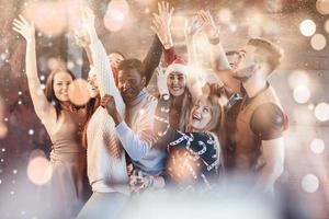 fiesta con amigos. les encanta la navidad. grupo de jóvenes alegres que llevan bengalas y flautas de champán bailando en la fiesta de año nuevo y luciendo felices. efecto suave de luz bokeh foto