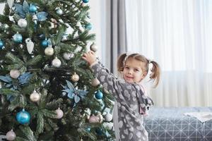 feliz navidad y felices fiestas. niña ayudando a decorar el árbol de navidad, sosteniendo algunos adornos navideños en la mano foto