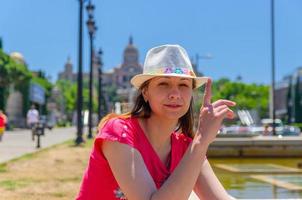 Young woman traveler with red dress and hat is sitting near fountain in Barcelona in sunny summer day