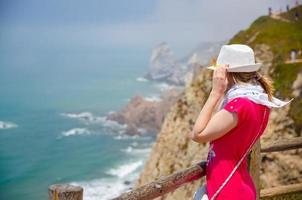 Young woman traveler with white hat and red dress staying near wooden fence at cliff of Cabo da Roca Cape Roca photo