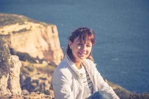Young beautiful girl traveler with white jacket looking at camera, squint and smile at Dingli Cliffs photo