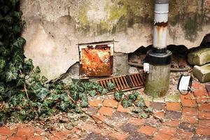 La hiedra inglesa se arrastra desde la esquina del edificio sobre el pavimento de baldosas rojas a lo largo de un muro de hormigón envejecido roto con una pequeña puerta oxidada y un tubo foto