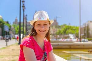 Young woman traveler with red dress and hat is sitting near fountain in Barcelona in sunny summer day photo