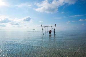 Lonely woman swinging on wooden swing over shallow and calm sea water with blue sky background photo