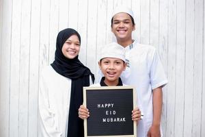 joven familia musulmana asiática con vestido tradicional y el niño que sostiene un tablero de cartas dice feliz eid mubarak foto