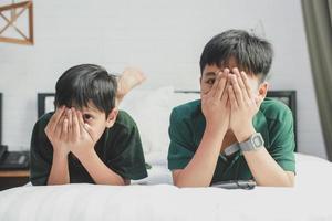dos hermanos acostados en la cama viendo la televisión con expresión de miedo cubriendo los ojos con las manos foto