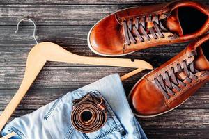 Brown leather casual shoes, jeans and belt on dark wooden background top view