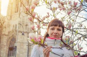 joven viajera con chaqueta blanca mirando a la cámara, sonriendo y tocando la rama de un árbol con flores en flor foto