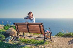 Young beautiful girl traveler with white jacket looking at camera, sit and smile on bench at Dingli Cliffs photo