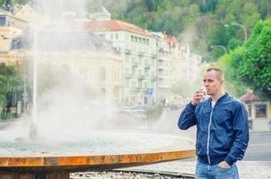 joven turista con chaqueta azul mira la cámara y bebe agua mineral terapéutica de la taza foto