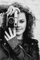 monochrome portrait of young beautiful cheerful woman with red curly hair taking pictures on film old fashioned camera on white bricks wall background. Film stylization photo