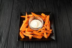 Fried sweet potatoes on a black plate with sauce on a wooden black background. photo