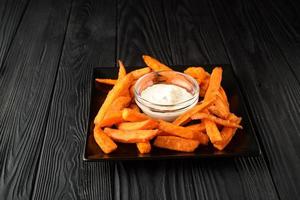 Fried sweet potatoes on a black plate with sauce on a wooden black background. photo