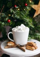 Coffee in a white cup with marshmallows. Morning festive coffee with traditional Italian cantuccini almond cookies. A cup of coffee on a background of green fir branches on a white stand. photo