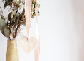 Light wooden heart pendant on a pink silk ribbon on a background of a golden shiny glittered vase with dried eucalyptus branches. photo