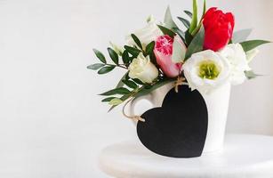 Black board heart copyspace. Spring bouquet in white vase on wooden white stand. Roses, tulips and lisianthus. photo