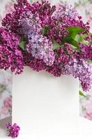 White sheet in beautiful blooming lilac on a white stand on a floral background. Greeting card, place for text, mock up photo