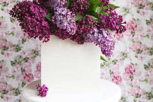 White sheet in beautiful blooming lilac on a white stand on a floral background. Greeting card, place for text, mock up photo