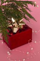 A fabulous gift in a red box with a gold ribbon under the Christmas tree, on a pink background. New Year's and Christmas. Copy space. photo