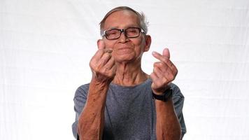 feliz homem idoso vestindo camiseta cinza fazendo mini sinal em forma de coração com as mãos no fundo branco do estúdio.