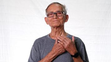 Happy elderly man wearing gray t-shirt holding hands on chest grateful heart on white studio background. video