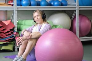 mujer rubia con auriculares descansando después de hacer ejercicio en un gimnasio. mujer sosteniendo una botella con agua y relajándose en el suelo después del entrenamiento físico. foto