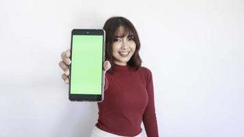hermosa chica asiática apunta en un teléfono de pantalla verde con un fondo borroso en la cara de fondo blanco foto