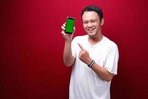 Smile Young Asian man pointing on green blank space on smartphone on red background. photo