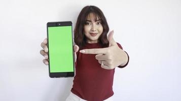 hermosa chica asiática apunta en un teléfono de pantalla verde con un fondo borroso en la cara de fondo blanco foto