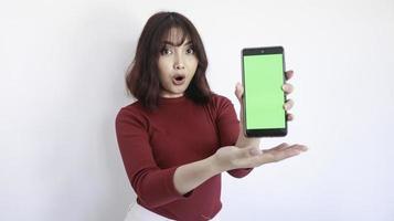 guau y conmociona a una hermosa chica asiática apuntando a un teléfono de pantalla verde con un fondo borroso en la cara en blanco foto