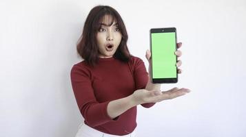 guau y conmociona a una hermosa chica asiática apuntando a un teléfono de pantalla verde con un fondo borroso en la cara en blanco foto