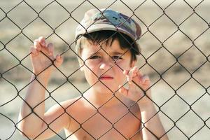 Sad child in a military cap behind a fence. Stop war concept photo