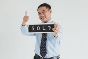 Smiling sales man wearing blue shirt and tie posing with thumb up and showing sold sign board at camera on white background photo