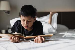 niño asiático acostado en la cama mientras lee un libro foto