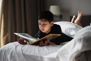 niño asiático acostado en la cama mientras lee un libro foto