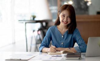 Cheerful young woman with laptop and jar and writing in notebook while sitting at table and working on freelance project at home