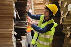 Female worker in safety uniform and hard hat, supervisor quality inspects packaging stock order supply at factory storage warehouse, piles of stacking paper manufacture, recycling production industry.