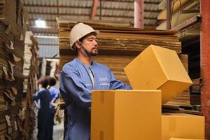 Asian male delivery worker in safety uniform and hard hat checks cardboard box, shipping orders at parcels warehouse, paper manufacture factory for the packaging industry, logistic transport service. photo