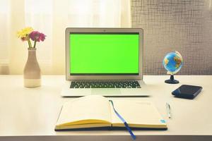 Green screen laptop computer sitting on a home work desk next to notebook, spring flowers and world globe stand. Concept minimalist digital student work space photo