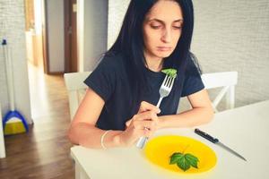 una mujer joven y atractiva come hojas verdes infelices en la cocina para perder peso. concepto de dieta desequilibrada y alimentos crudos poco saludables foto